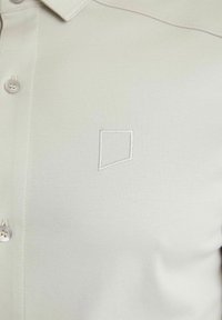 Chemise beige clair à boutons avec un petit logo géométrique brodé sur le côté gauche de la poitrine, près des boutons.