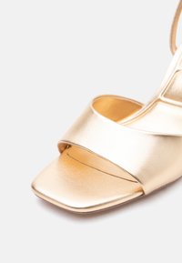 Sandale aurii cu vârf decupat, cu o baretă largă în față și vârf pătrat, pe fundal alb.