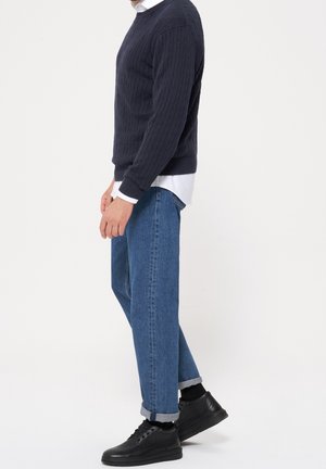 Personne portant un pull en maille torsadée bleu marine par-dessus une chemise blanche, un jean bleu retroussé, des chaussettes noires et des baskets en cuir noires, debout de profil.