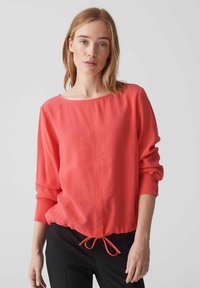 Blusa a maniche lunghe color corallo con scollatura ampia, orlo elastico arricciato e polsini con bottoni, realizzata in tessuto leggero e morbido.