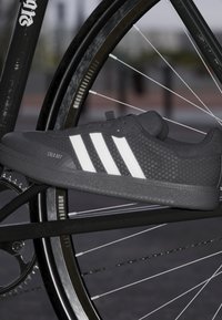 Czarny sneaker z teksturowaną cholewką, białym wzorem w trzy paski, płaską gumową podeszwą i wzmocnionym przodem. Ułożony w pobliżu koła rowerowego.