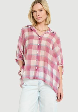 Žena s blond bobmi v voľnej ružovo-bielej károvanej košeli na gombíky a svetlo-modrých džínsoch, stojaca s rukami v kapse na sivom pozadí.