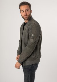 Giacca bomber in suede grigio con polsini e orlo a coste, dotata di zip frontale, tasche laterali e una toppa sulla manica sinistra.