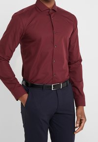 Bordeaux langemouwen overhemd met een button-down kraag en donkere knopen, gecombineerd met donkere broek en een zwarte riem met zilveren gesp.