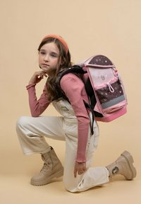 Zaino con tessuto rosa e marrone, motivi a stella, spallacci regolabili e dettagli in metallo. Il modello indossa stivali beige e una salopette crema.