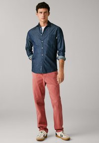 Camicia di denim blu scuro con bottoni sul davanti e maniche arrotolate, abbinata a pantaloni rosso chiaro e sneaker casual con dettagli.