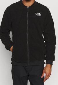 Homme portant une veste polaire noire zippée avec le logo The North Face sur le côté gauche de la poitrine, les mains partiellement dans les poches.