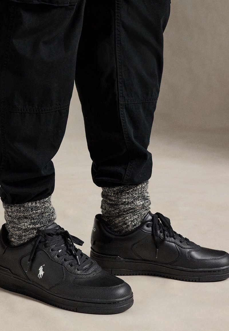 Baskets basses noires avec lacets, logo blanc sur le côté, portées avec des chaussettes épaisses grises à motifs et un pantalon noir sur un fond uni.