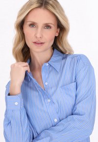 Camicia button-up azzurro chiaro con righe bianche verticali, maniche lunghe, colletto e taschino sul petto. Il tessuto sembra liscio e traspirante.