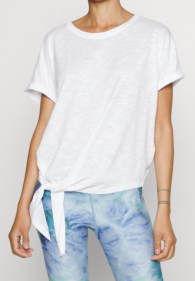 Camiseta blanca de manga corta con detalle frontal anudado, hecha de un material ligero y texturizado, combinada con leggings marmoleados en azul y verde.