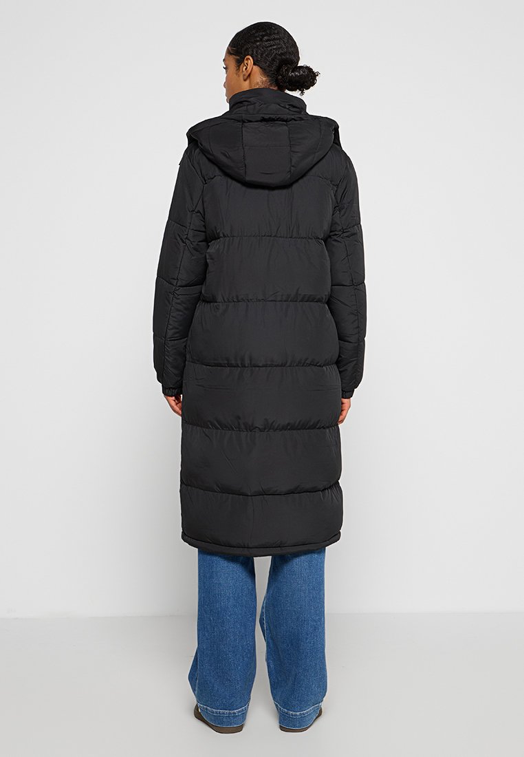 Zwarte gewatteerde jas met capuchon, met doorgestikte delen en een lange silhouet, gecombineerd met wijde blauwe jeans. Gladde textuur.