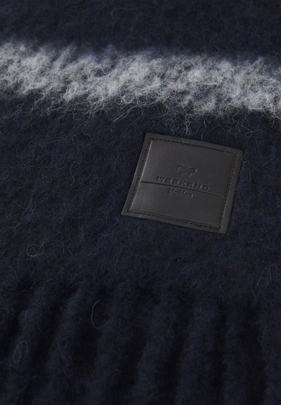 Tissu en maille bleu marine avec une texture douce et duveteuse, présentant une rayure diagonale blanche. Comprend une étiquette en cuir noir avec l'inscription "WEEKEND" embossée.