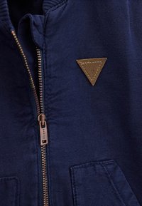 Hoodie zippé bleu marine en tissu texturé, doté d'un patch en cuir marron en forme de triangle et d'une fermeture à double zip.