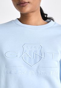 Világoskék pulóver bordás kerek nyakkal, domború GANT logóval és pajzs mintával a mellkason. Puha szövettextúra.
