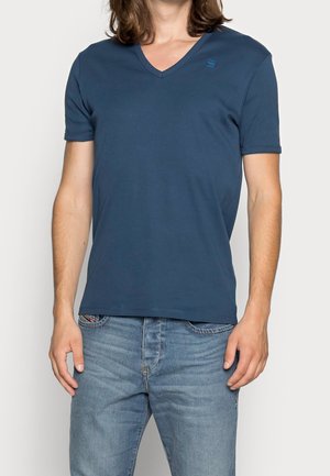 Navyblaues V-Ausschnitt T-Shirt aus weichem Stoff mit kurzen Ärmeln, mit einem dezenten Logo auf der Brust. Kombiniert mit hellblauen Jeans.