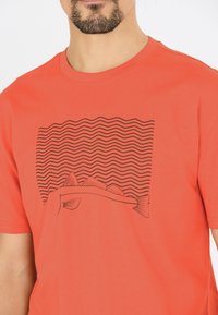 Röd bomullströja med en svart linjediagram av vågor och en fisk. Korta ärmar och rundad halsringning. Slät textur.