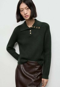 Mujer con cabello lacio y oscuro, vistiendo un suéter ribeteado verde oscuro con botones dorados y una falda de cuero marrón oscuro, frente a un fondo liso.