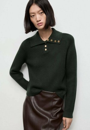 Mujer con cabello lacio y oscuro, vistiendo un suéter ribeteado verde oscuro con botones dorados y una falda de cuero marrón oscuro, frente a un fondo liso.
