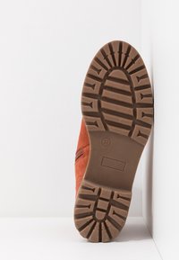 Semelle d'une botte marron à semelle en caoutchouc avec des crampons profonds, taille 37, reposant verticalement contre un mur blanc sur une surface claire.