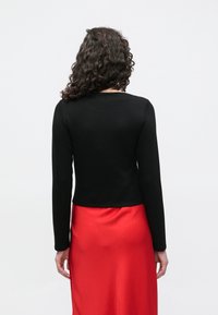 Μαύρο μακρυμάνικο τοπ σε εφαρμοστή γραμμή, με στρογγυλή λαιμόκοψη, συνδυασμένο με μια λεία, κόκκινη σατέν φούστα με ελαφριά λάμψη.