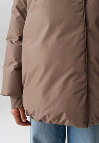 Giacca puffo marrone con una finitura liscia e lucida, dotata di zip frontale, polsini elastici e vestibilità morbida. Mano appoggiata di lato.