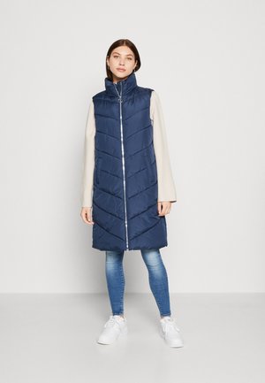 Veste doudoune bleu marine avec un col haut, des coutures en chevron vertical, une fermeture éclair à l'avant et des poches latérales, associée à un jean bleu clair et des baskets blanches.