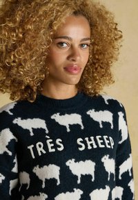 Frau mit lockigem blonden Haar trägt einen dunkelblauen Pullover mit einem Muster aus weißen Schafen und dem Text "TRÈS SHEEP" auf einem beigen Hintergrund.