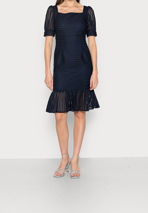 Femme portant une robe en dentelle bleu marine ajustée avec des manches bouffantes, un décolleté carré et un ourlet volanté, associée à des talons bleu clair à brides.