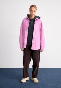 Ružová kordurová košeľa-jacket vrstvená nad tmavým mikinou, spárovaná so hnedými kordurovými nohavicami a béžovými teniskami na modrej kobercovej podlahe.