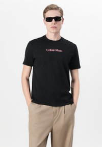 Black cotton T-shirt with a round neckline, featuring a red and white "Calvin Klein" logo on the front. Worn with beige trousers and sunglasses.