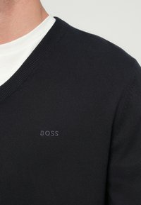 Czarny sweter z dekoltem w serek wykonany z drobno dzianinowej tkaniny, z białym wewnętrznym kołnierzem i subtelnym haftowanym logo "BOSS" na piersi.