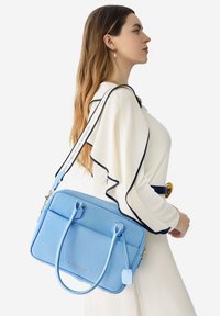 Bolso de mano de cuero azul claro con dos asas superiores y una correa ajustable. Presenta una textura suave, herrajes en tono dorado y detalle del logotipo.