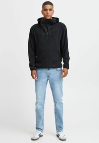 Svart hettegenser med glidelås i halsen, snorhette og frontlommer, kombinert med lyseblå jeans og hvite joggesko.