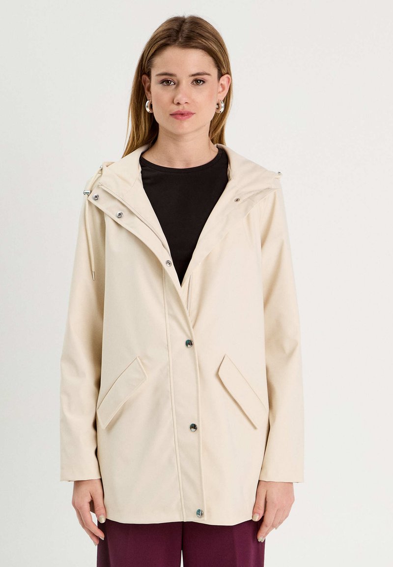 Veste beige, résistante à l'eau, avec capuche, dotée d'une fermeture à boutons, de poches latérales et d'une texture lisse. Design simple avec des accents minimaux.