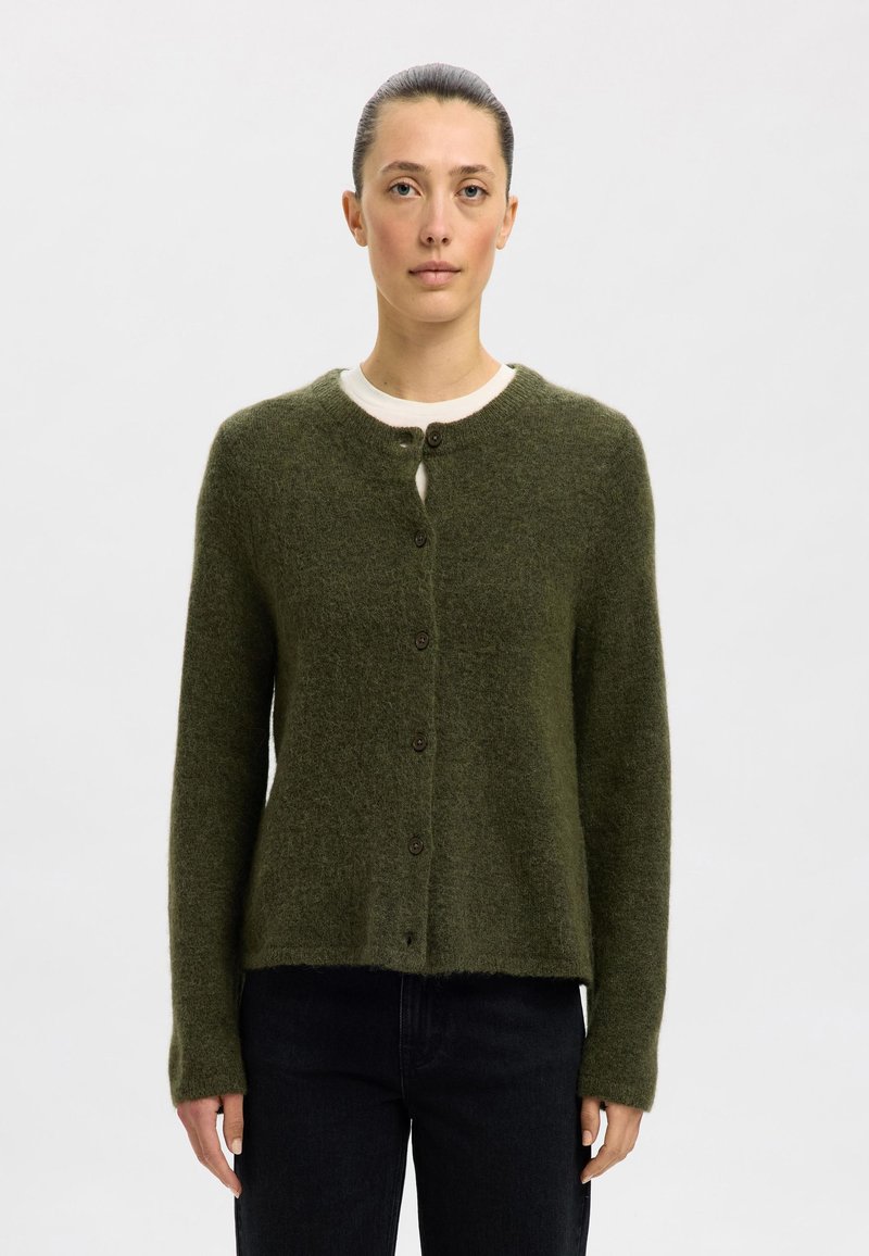 Πράσινο, υφασματοποιημένο κάρντιγκαν από μαλακό υλικό, με στρογγυλή λαιμόκοψη και καφέ κουμπιά, τοποθετημένο πάνω από ένα λευκό πουκάμισο.