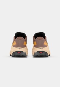 Et par brune og tan sportsko med sorte såler og gule aksenter, sett fra baksiden på en ensfarget bakgrunn.