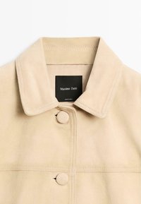 Giacca in suede beige con ampio colletto, due bottoni coperti, cuciture visibili e un'etichetta nera interna con la scritta "Massimo Dutti".