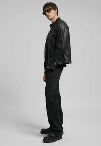 Zwarte leren jas met een rits aan de voorkant, gecombineerd met zwarte jeans en stevige zwarte laarzen. Model draagt zwarte zonnebril en staat opzij geposeerd.