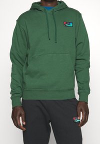 Grön huva tröja gjord av mjukt tyg med en kenguruficka. Har ett flerfärgat Nike-logopatch på bröstet.