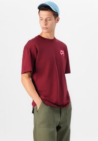 T-shirt en coton bordeaux avec des manches courtes et un logo Nike rose. Associé à un pantalon vert olive et une casquette bleu clair.