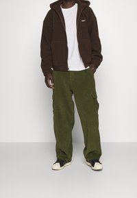 Person klädd i en brun zip-up hoodie, vit t-shirt, olivgröna cargobyxor och svart-vita sneakers mot en enhetlig bakgrund.