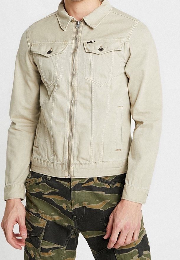 Mężczyzna w beżowej kurtce na suwak z zapinanymi na guziki kieszeniami na piersi oraz w kamuflażowych spodniach typu cargo, stojący na tle jednolitego tła.