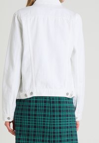 Femme portant une veste en denim blanc sur une jupe à carreaux vert et bleu marine, vue de dos sur un fond uni.