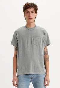 Grå bomulds T-shirt med afslappet pasform, korte ærmer og en venstre brystlomme med et rødt mærke. Båret med blå jeans.