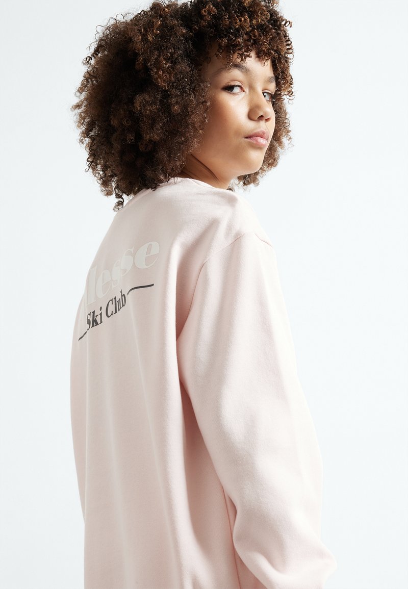 Sudadera de color rosa claro con un ajuste relajado, que presenta un diseño gráfico en blanco y negro en la parte posterior que dice "Ski Club". Textura suave.