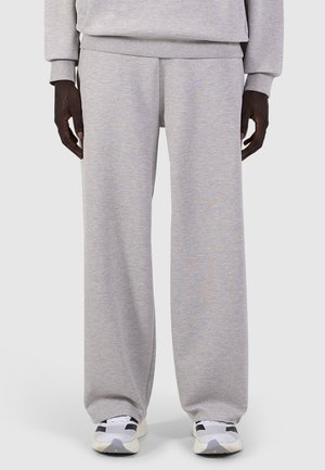 Persoon die lichtgrijze wijde joggingbroek en witte sneakers draagt, staand met ontspannen handen langs de zij.