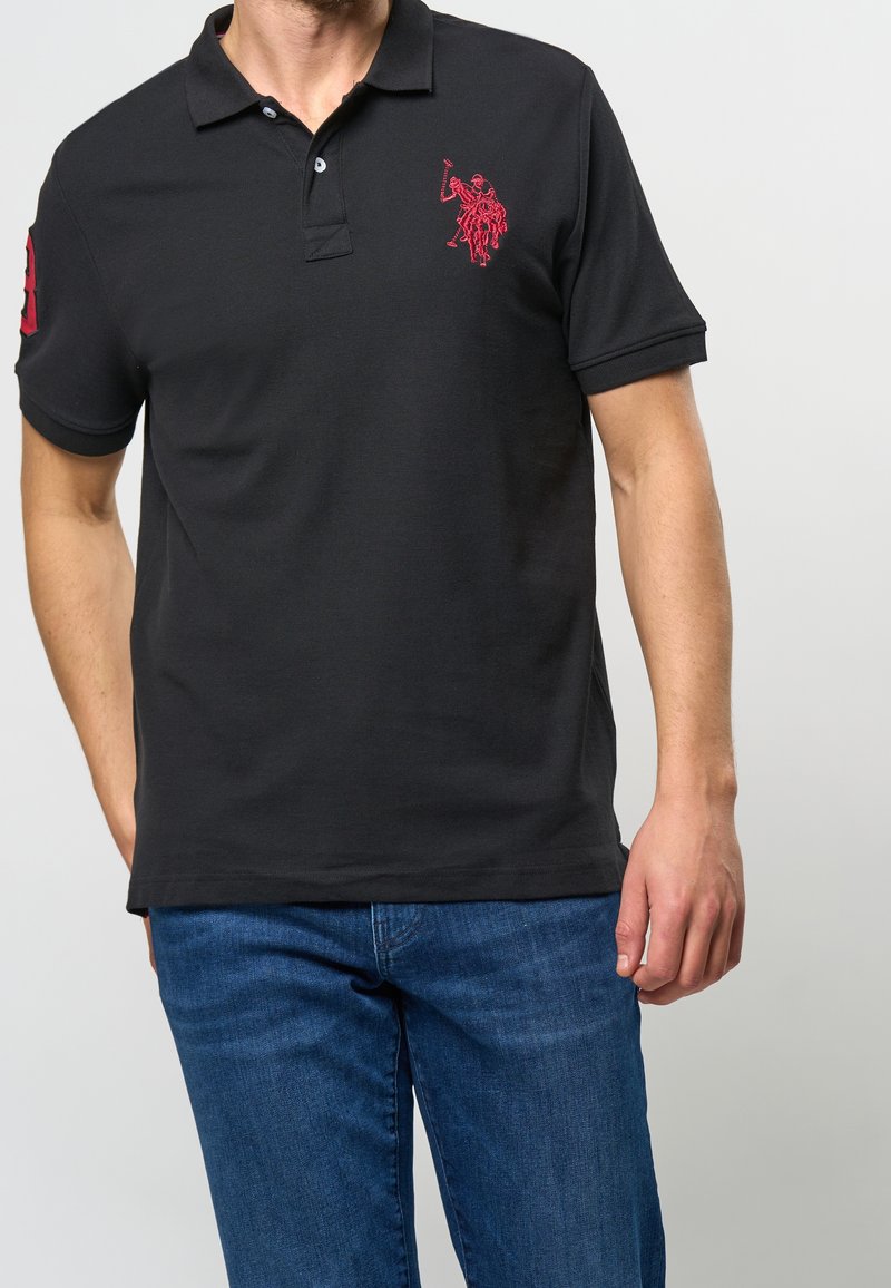 Homme portant un polo noir avec un logo brodé en rouge d'un joueur de polo sur la poitrine et un écusson numéroté rouge sur la manche, assorti à un jean bleu.