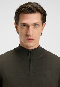 Donkergroene, geribbelde gebreide pullover met een hoge kraag en half-ritssluiting. De gladde textuur en de aansluitende pasvorm versterken de moderne uitstraling.