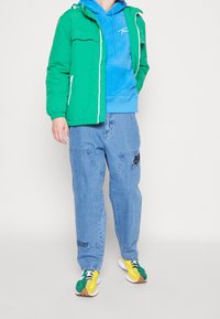 Giacca verde trapuntata indossata sopra un hoodie blu, abbinata a pantaloni in denim azzurro chiaro con ricami neri e sneakers multicolori.