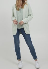 Cardigan lavorato a maglia di un verde chiaro con tasche, abbinato a una blusa mint a pois e jeans blu. Modella con scarpe da ginnastica bianche.